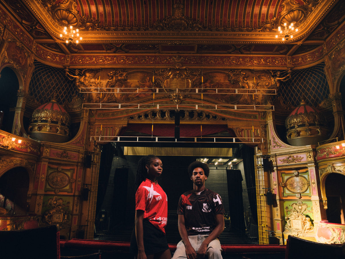 Two Hackney Women's FC players in red and black Hackney Empire jerseys on the Hackney Empire stage